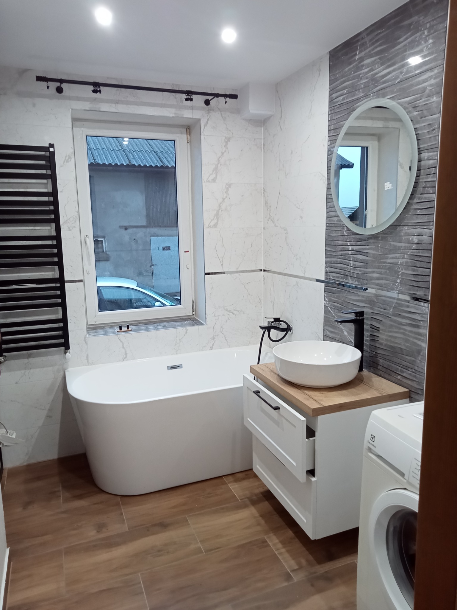 Nowoczesna łazienka z wolnostojącą wanną, umywalką nablatową na szafce z otwartą szufladą, grzejnikiem drabinkowym i pralką Electrolux, wykończona płytkami imitującymi marmur i drewno.