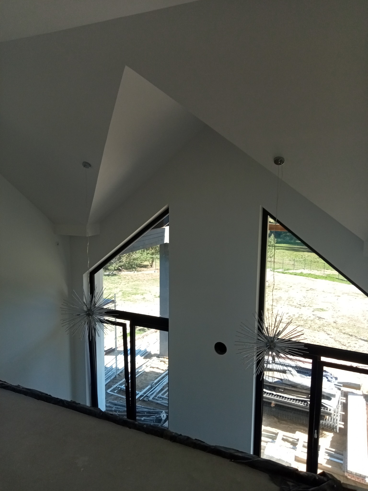 Wnętrze domu w trakcie wykańczania z dwoma trójkątnymi oknami i dekoracyjnymi lampami w kształcie jeża, widoczne materiały budowlane na zewnątrz.