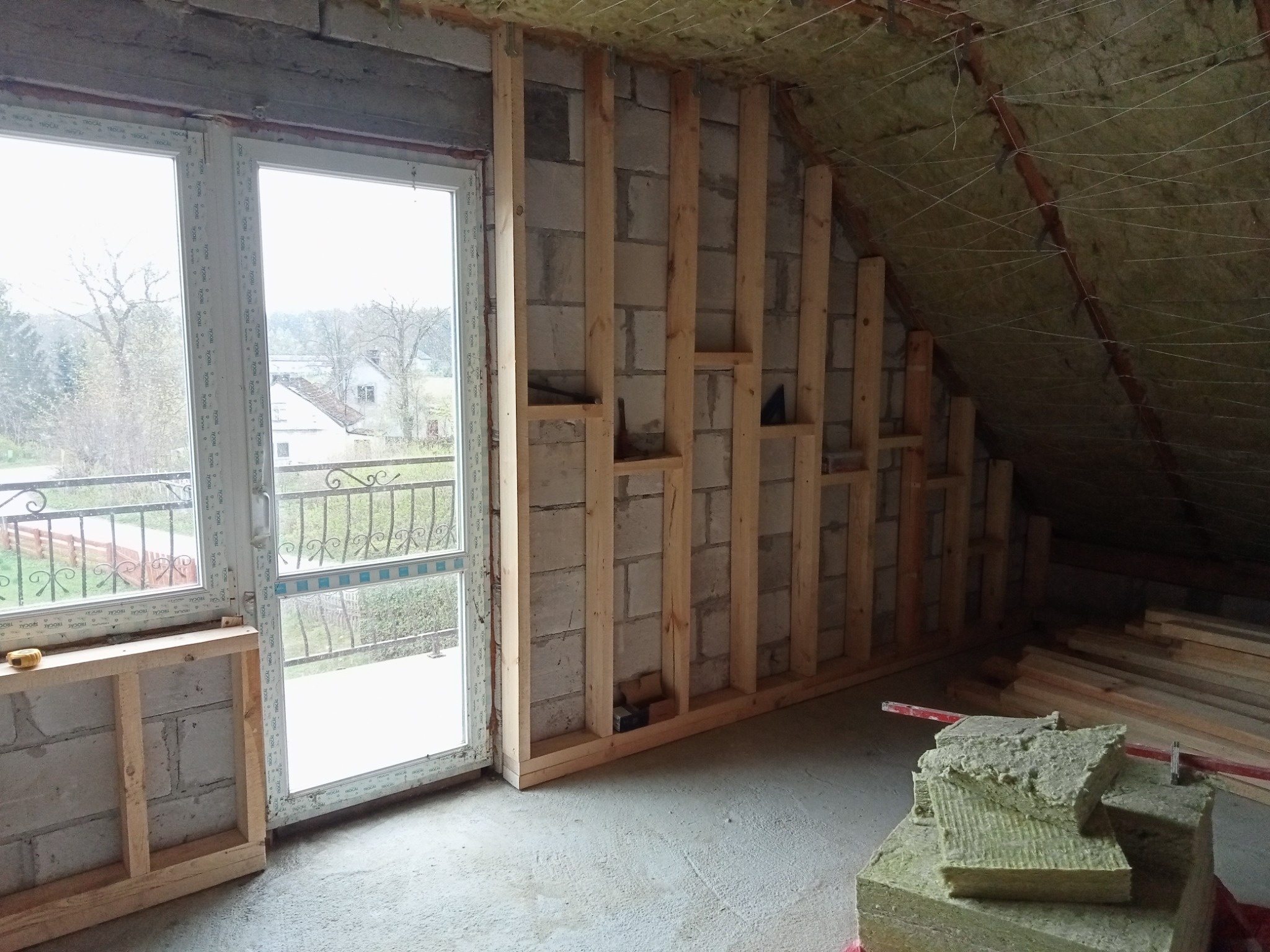 Poddasze w trakcie adaptacji: widoczna konstrukcja drewniana ścianki działowej z izolacją wełną mineralną oraz okno balkonowe z widokiem na zewnątrz.