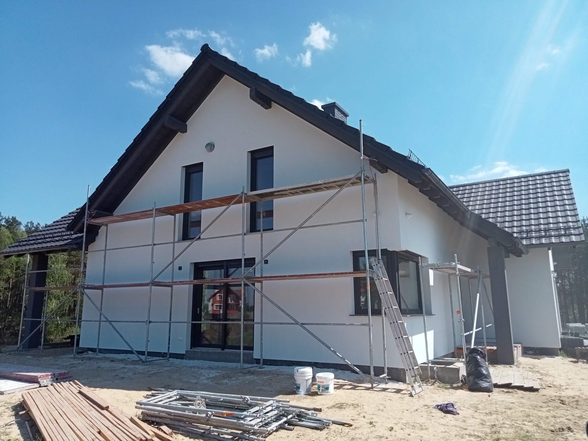 Nowoczesny dom jednorodzinny z jasną elewacją, w trakcie malowania, z widocznymi rusztowaniami i materiałami budowlanymi na ziemi.