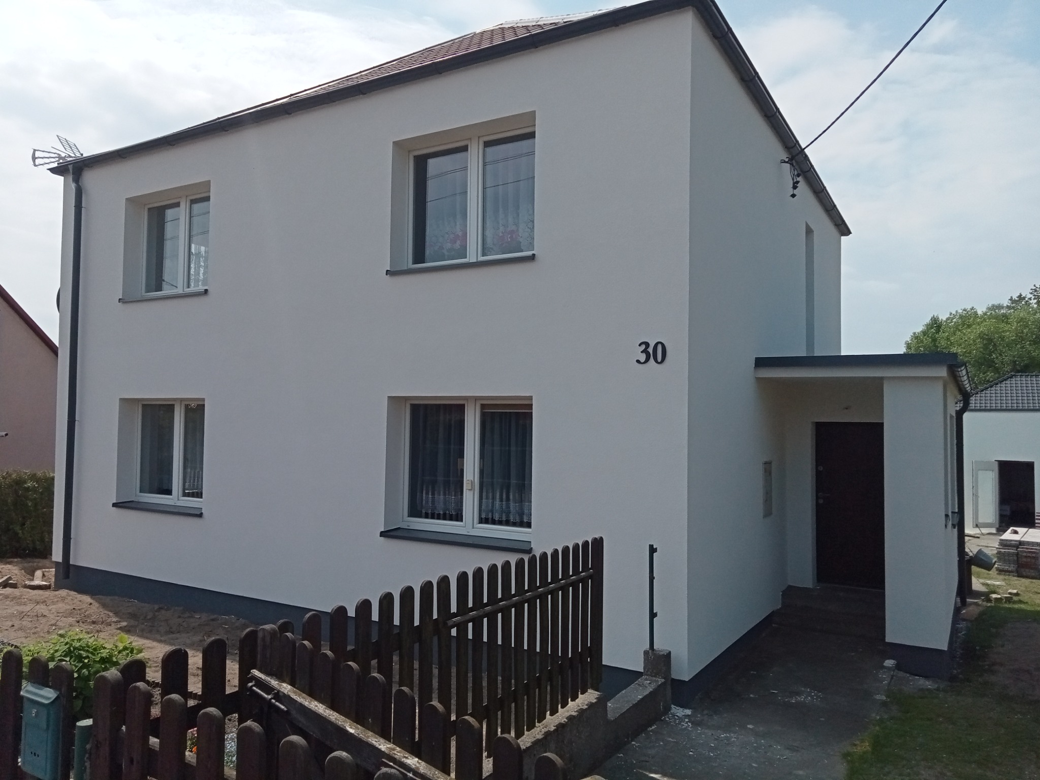 Dwupiętrowy dom z jasną, gładką elewacją, numerem 30 na ścianie i brązowym ogrodzeniem w pierwszym planie, widoczne okna z firankami i zadaszone wejście.