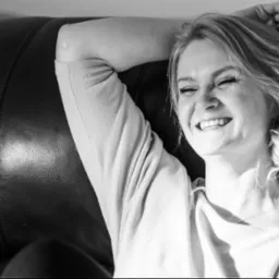 Czarno-białe zdjęcie roześmianej kobiety z włosami upiętymi w luźny kok, leżącej na skórzanej sofie, z ręką pod głową, wpadające światło podkreśla strukturę skóry.
