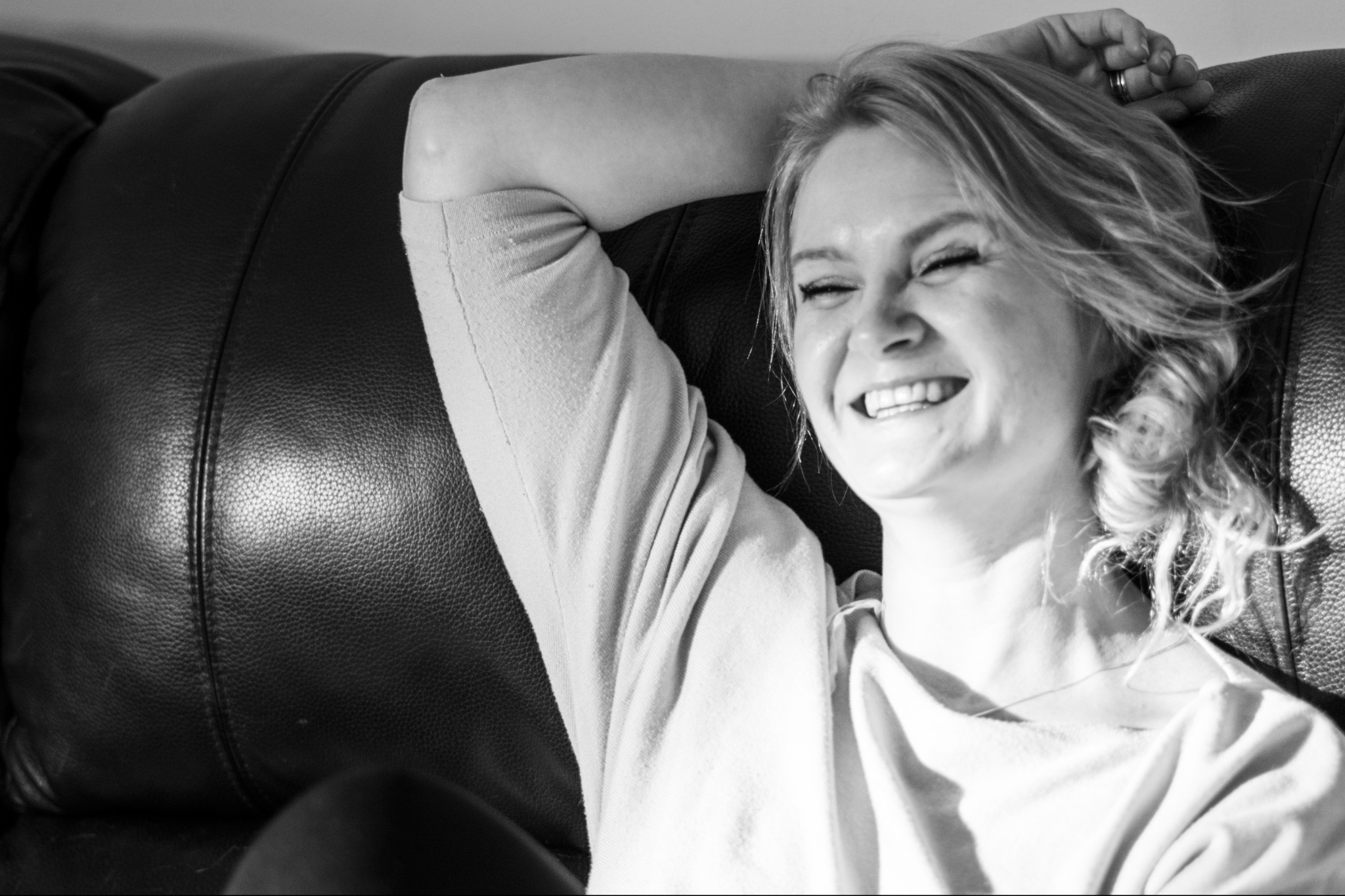 Czarno-białe zdjęcie roześmianej kobiety z włosami upiętymi w luźny kok, leżącej na skórzanej sofie, z ręką pod głową, wpadające światło podkreśla strukturę skóry.