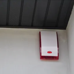 Zewnętrzna sygnalizacja alarmowa z czerwoną obudową, zamontowana na ścianie budynku w Szubinie, widoczna pod ciemną podbitką dachu.