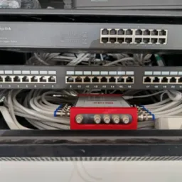 Widok z bliska na szafę rack z zamontowanym switchem TP-Link, panelem krosowniczym oraz czerwonym rozdzielaczem sygnału Grab, z licznymi kablami sieciowymi.