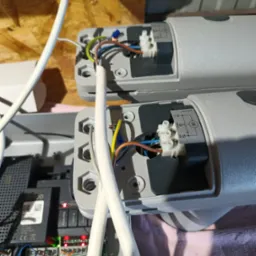 Szczegółowe ujęcie podłączenia przewodów elektrycznych do siłowników bramy automatycznej, widoczne złącza, kable różnego koloru oraz schemat elektryczny na obudowie.