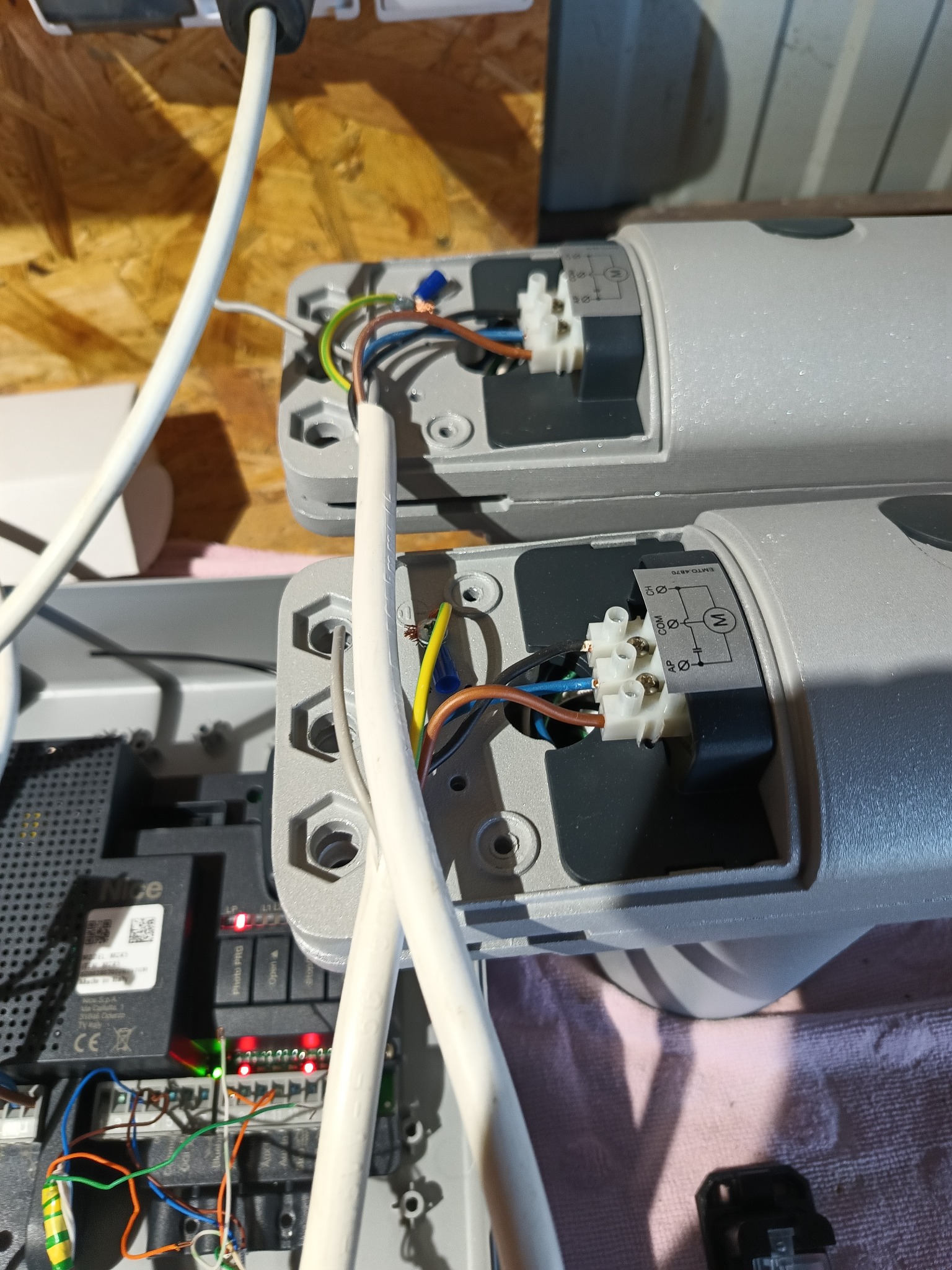 Szczegółowe ujęcie podłączenia przewodów elektrycznych do siłowników bramy automatycznej, widoczne złącza, kable różnego koloru oraz schemat elektryczny na obudowie.