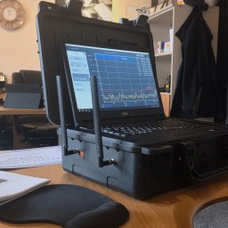 Agencja Detektywistyczna Biuro bezpieczeństwa SPECTRUM & Detektyw NOWICKI - Analizator widma radiowego Fujitsu w czarnej obudowie, z antenami, na drewnianym biurku. Na ekranie wykres fal radiowych. Obok podkładka pod mysz i certyfikat.