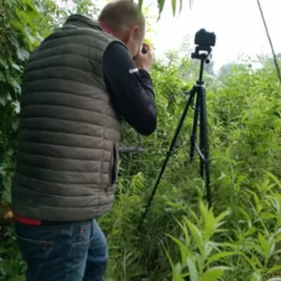 Mężczyzna w kamizelce i kaloszach robi zdjęcie aparatem na statywie w zaroślach.