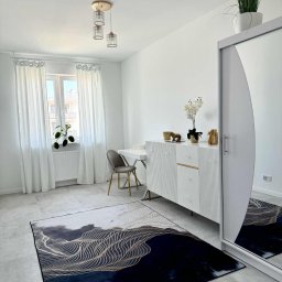 R-Style usługi remontowo-wykończeniowe Rafał Wyrzykowski - Jasne wnętrze pokoju z białymi ścianami, szarymi płytkami podłogowymi i dywanem w odcieniach szarości i granatu. Widoczne białe meble, okno z białymi zasłonami i dekoracje.