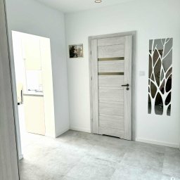 R-Style usługi remontowo-wykończeniowe Rafał Wyrzykowski - Jasne wnętrze: szare płytki podłogowe, białe ściany z dekoracją drzewa, drzwi w odcieniach szarości i widok na kuchnię. Nowoczesne oświetlenie sufitowe.