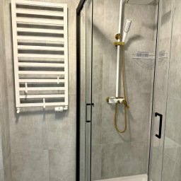 R-Style usługi remontowo-wykończeniowe Rafał Wyrzykowski - Nowoczesna kabina prysznicowa z czarną ramą, biała armatura z złotymi akcentami i grzejnik drabinkowy na szarej, płytkowej ścianie. Minimalistyczny design.