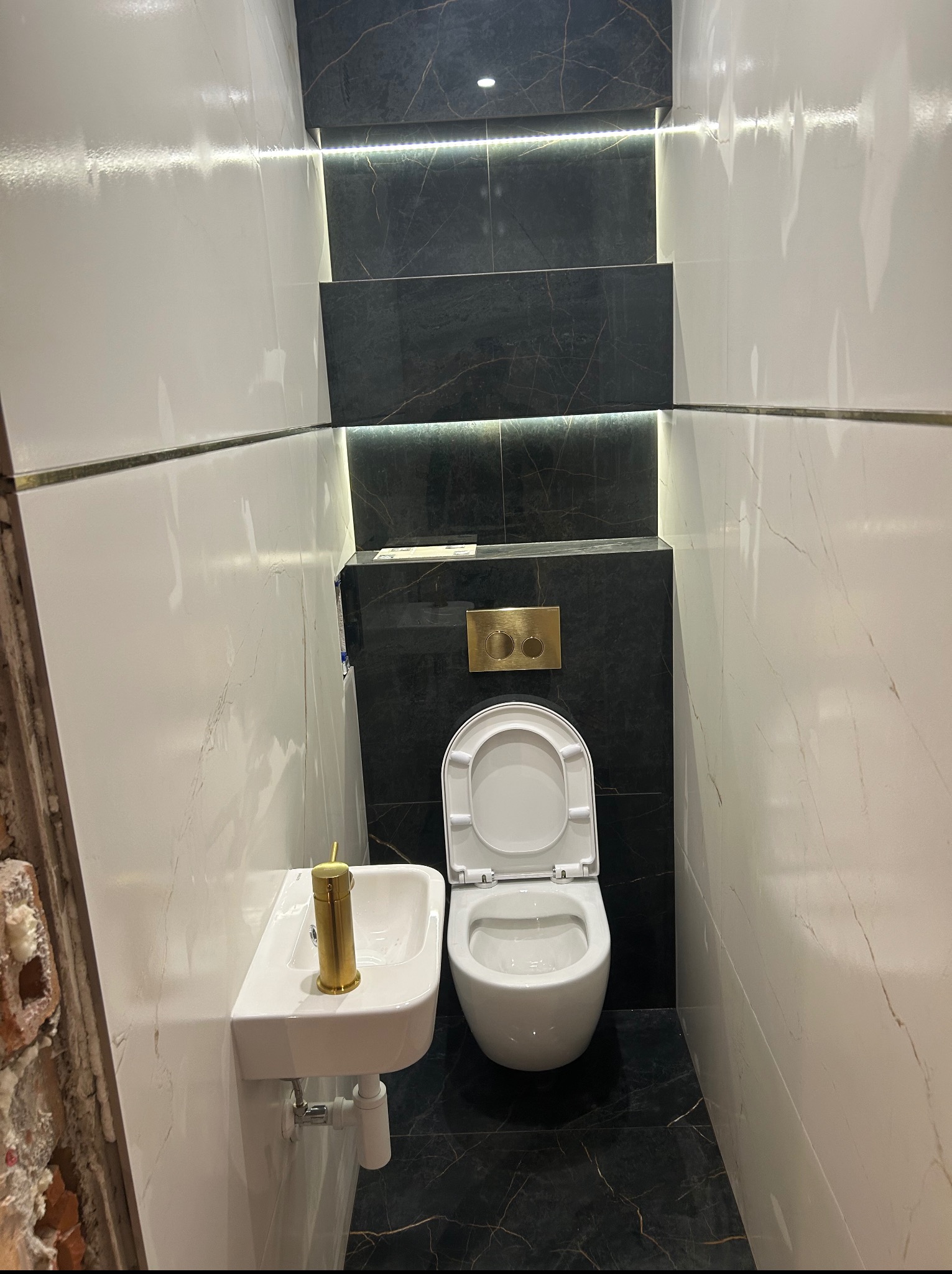 Wykończona łazienka w bieli i czerni z marmurkowym wzorem, podświetleniem LED i złotą armaturą. Umywalka narożna i podwieszana toaleta.