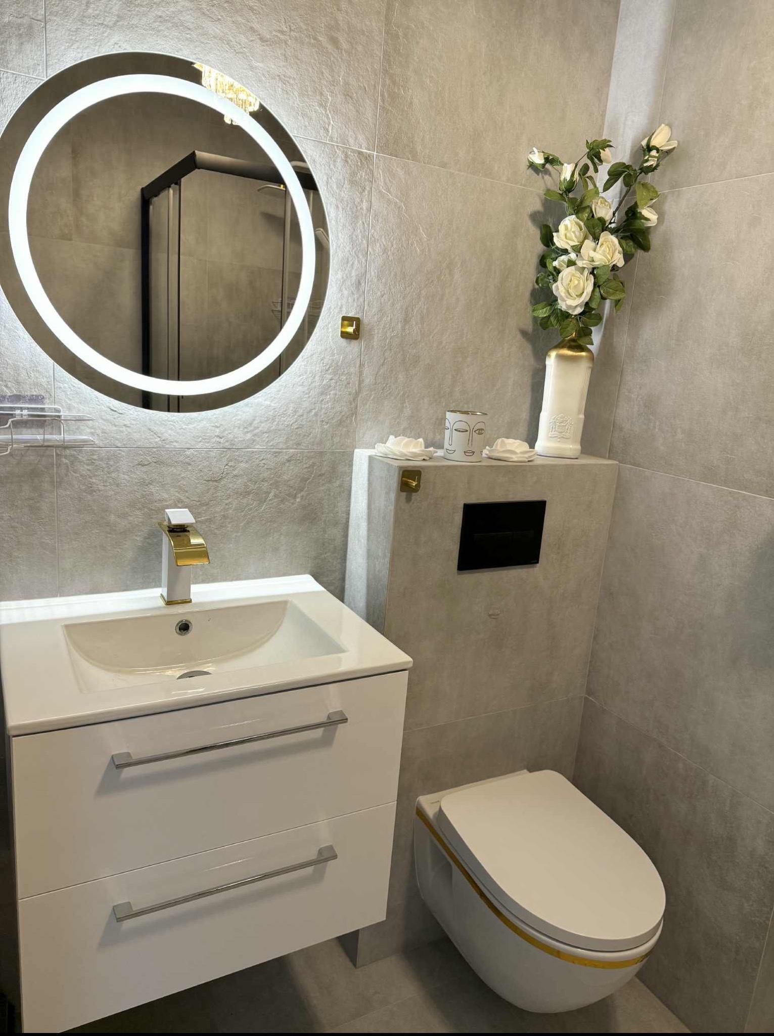 Nowoczesna łazienka z szarymi płytkami, okrągłym lustrem z podświetleniem LED, białą szafką z umywalką i złotą armaturą oraz podwieszaną toaletą.