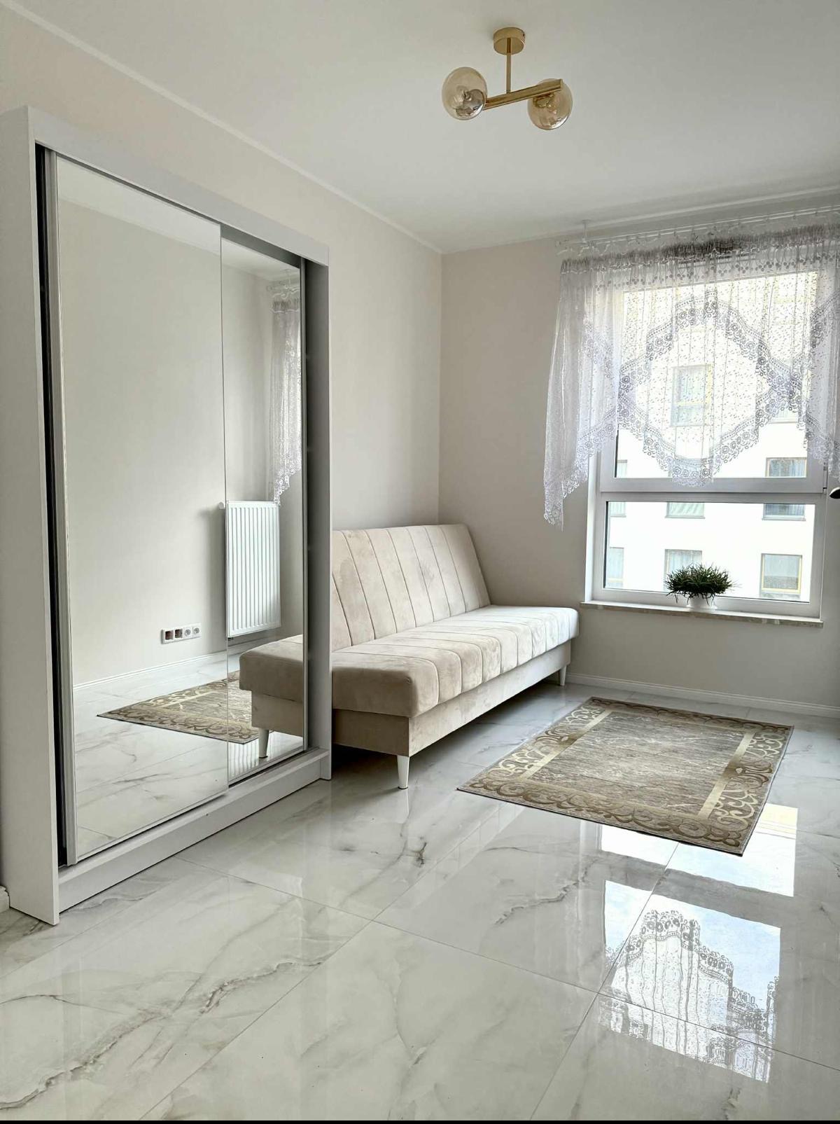 Jasne wnętrze pokoju z marmurową podłogą, beżową sofą i szafą z lustrem. Okno z koronkową firanką i dywan dodają przytulności. Nowoczesne oświetlenie sufitowe.