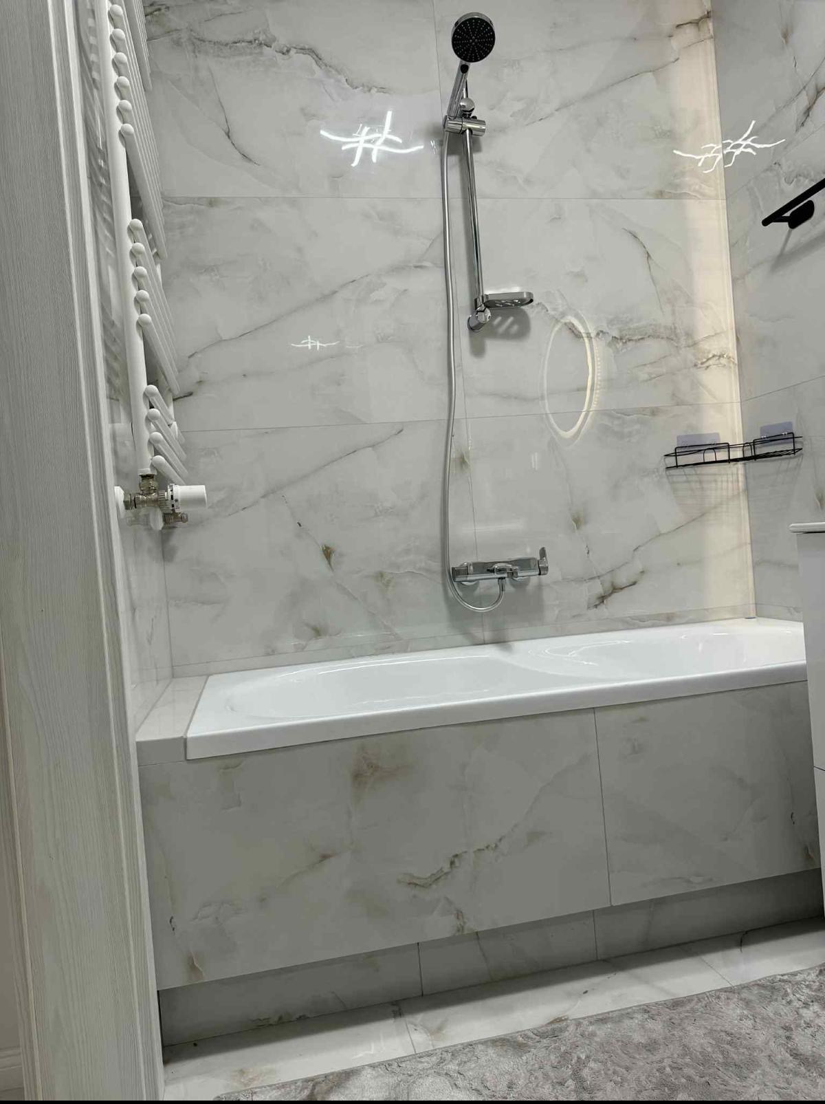 Wykończona łazienka z wanną obudowaną płytkami imitującymi marmur, chromowaną armaturą i grzejnikiem drabinkowym. Widoczne detale wykończenia.