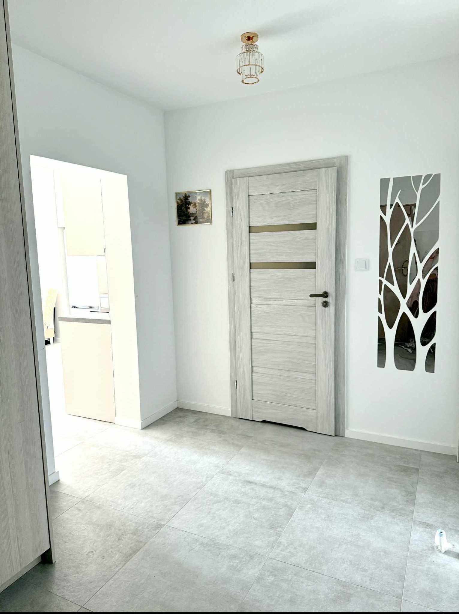 Jasne wnętrze: szare płytki podłogowe, białe ściany z dekoracją drzewa, drzwi w odcieniach szarości i widok na kuchnię. Nowoczesne oświetlenie sufitowe.