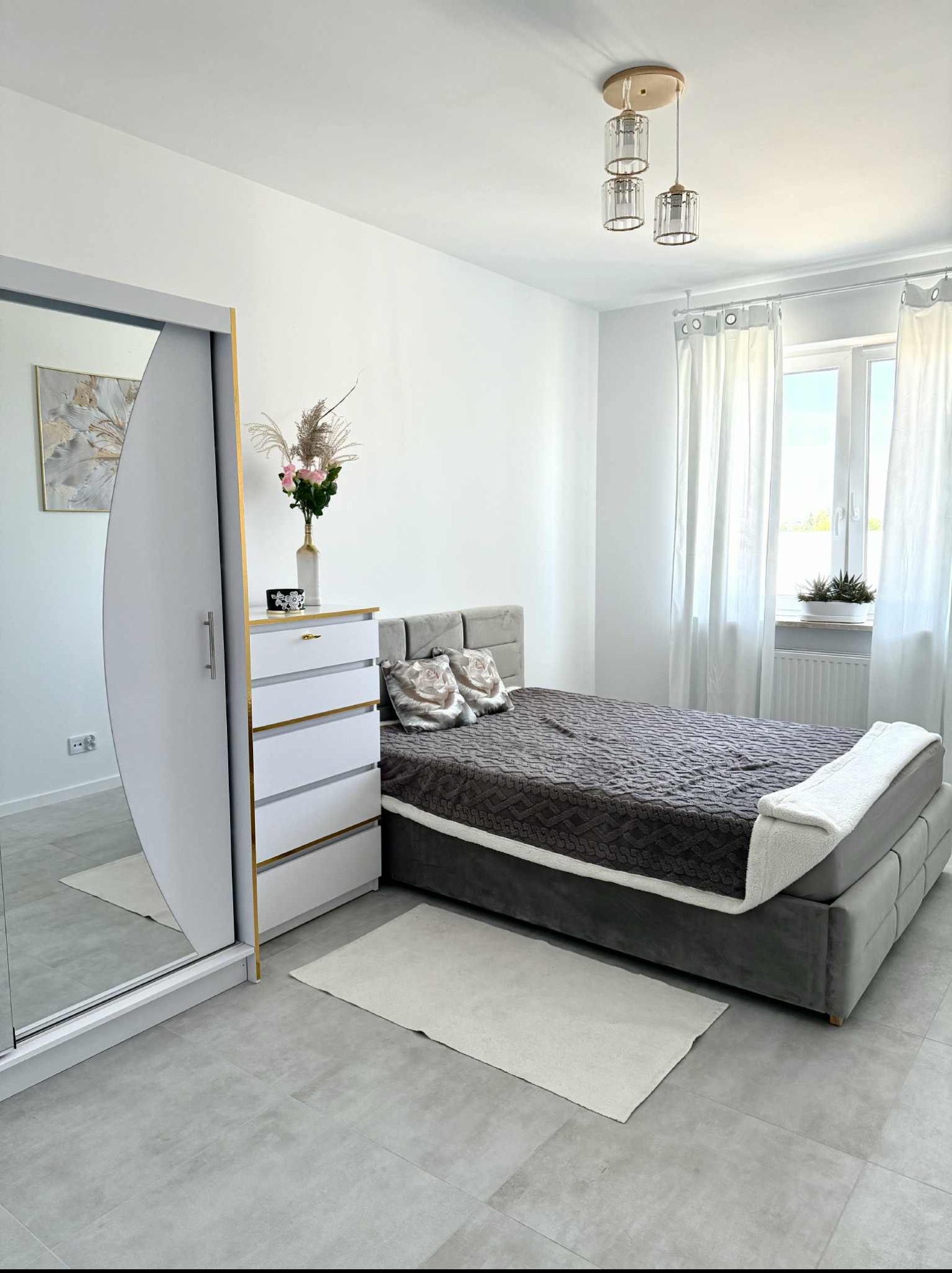 Jasna sypialnia z szarym łóżkiem, przesuwną szafą z lustrem i kwiatami w wazonie. Minimalistyczny design, szare płytki podłogowe i naturalne światło z okna.
