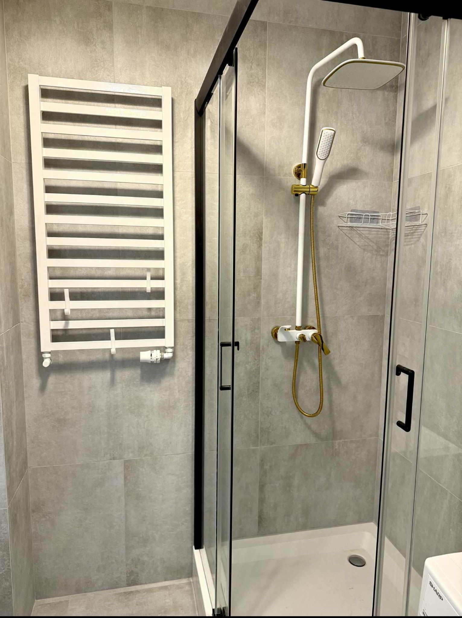 Nowoczesna kabina prysznicowa z czarną ramą, biała armatura z złotymi akcentami i grzejnik drabinkowy na szarej, płytkowej ścianie. Minimalistyczny design.