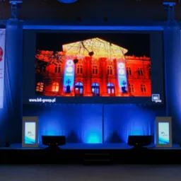 Scena z dużym ekranem LED wyświetlającym budynek w czerwonej iluminacji, boczne banery z logo Toyota Motor Europe, niebieskie oświetlenie sceniczne i dwa monitory na podeście.