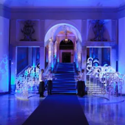 Uroczyste wejście do sali balowej z niebieskim oświetleniem, dekoracjami z liczb, projekcjami logo Totalizatora Sportowego i obsługą kelnerską.