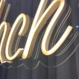 Fragment neonowego napisu w ozdobnej czcionce, podświetlonego ciepłym światłem, na ciemnym tle.