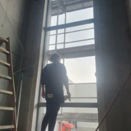 Pracownica z tyłu, w ciemnych ubraniach, myje okno w surowym wnętrzu, widoczny skrobak, drabina i czerwona ciężarówka za oknem.