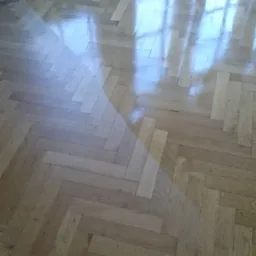 Parkiet ułożony w jodełkę, świeżo po polerowaniu, z widocznym odbiciem okna i jasnym oświetleniem.