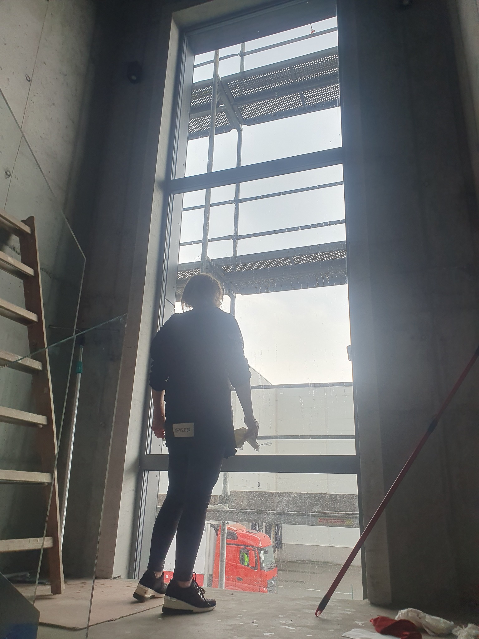 Pracownica z tyłu, w ciemnych ubraniach, myje okno w surowym wnętrzu, widoczny skrobak, drabina i czerwona ciężarówka za oknem.