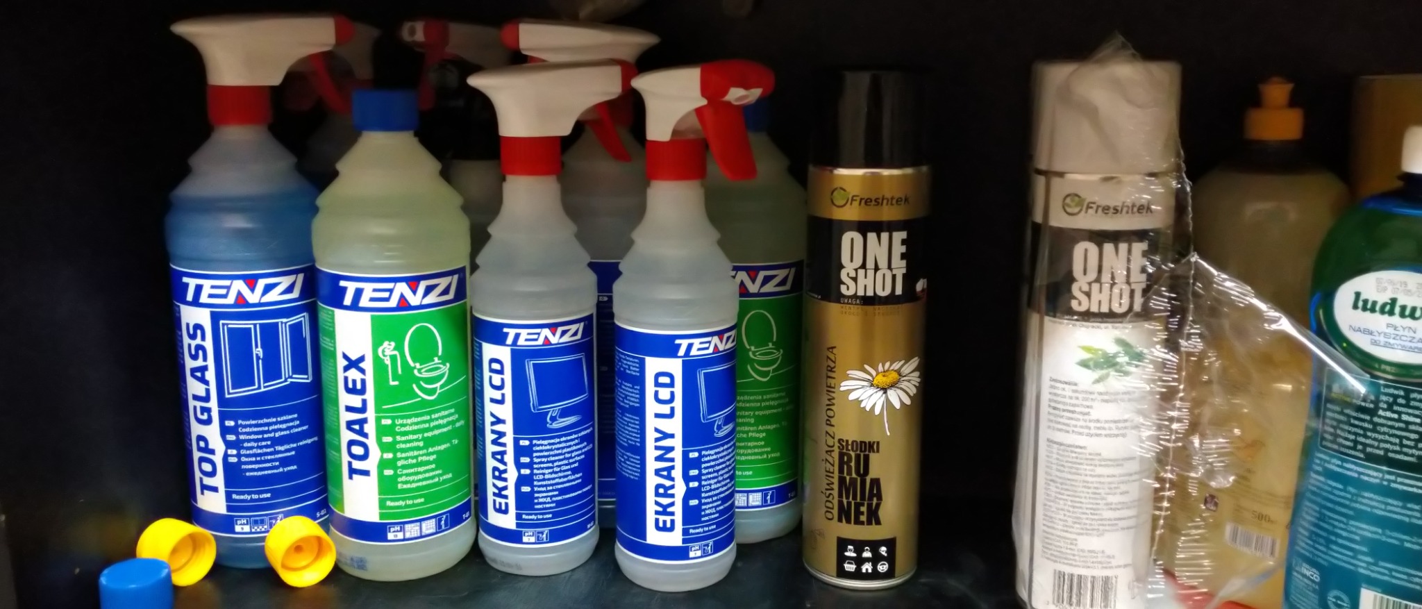 Półka z różnorodnymi środkami czyszczącymi marki Tenzi, w tym płyny do szyb, toalet i ekranów LCD, oraz odświeżacze powietrza Freshtek One Shot, ustawione w rzędzie w ciemnym pomieszczeniu.