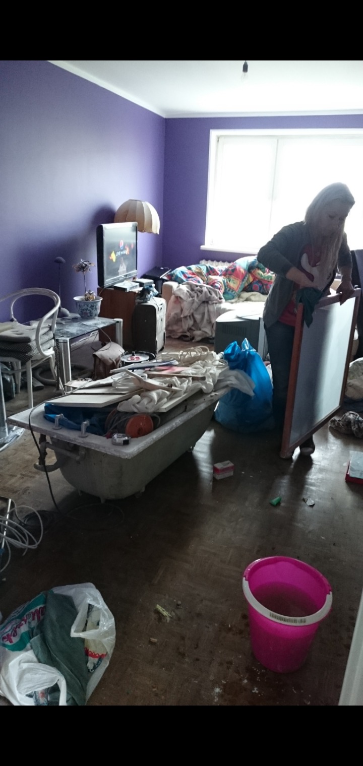 Pokój w trakcie opróżniania: wanna wypełniona przedmiotami, kobieta czyści dużą ramę, widoczny bałagan i wiadro z wodą.