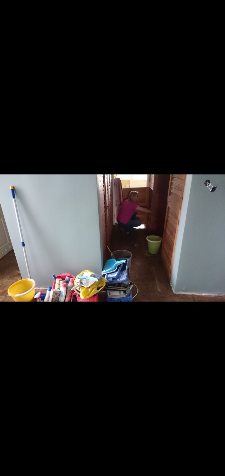 Kobieta w kucki czyści podłogę w wąskim pomieszczeniu z drewnianymi drzwiami, widoczne środki czystości i wiaderka.