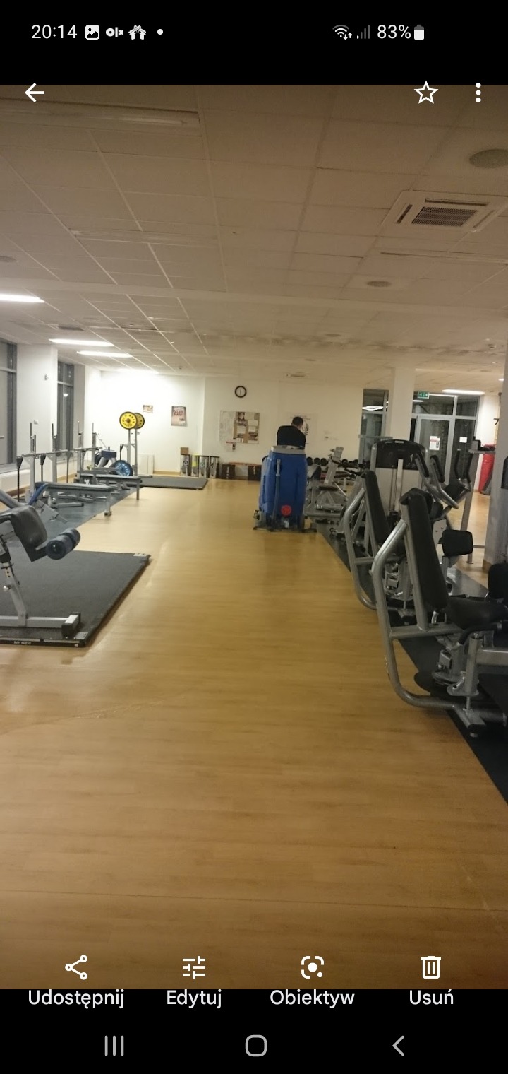 Maszyna do czyszczenia podłóg obsługiwana przez pracownika w tle sali gimnastycznej z wyposażeniem do ćwiczeń.