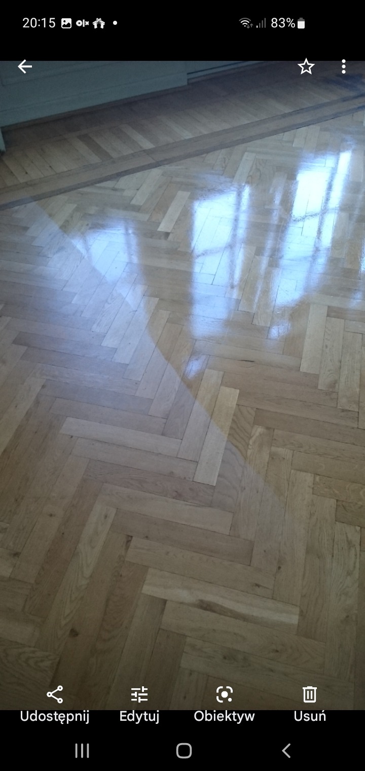 Błyszczący parkiet ułożony w jodełkę, odbijający światło okna, widoczne ślady po remoncie.