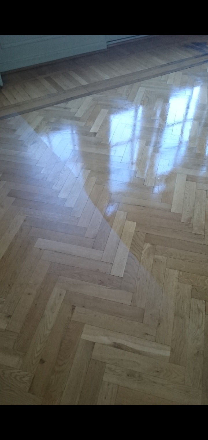 Parkiet ułożony w jodełkę, świeżo po polerowaniu, z widocznym odbiciem okna i jasnym oświetleniem.