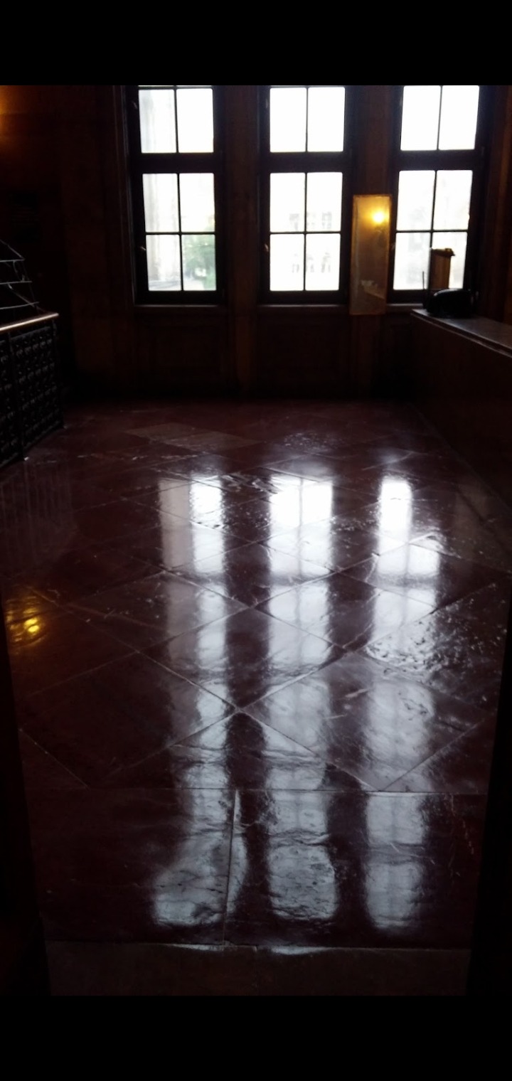 Błyszcząca, mokra podłoga z płytek w ciemnym pomieszczeniu z oknami, odbijająca światło dzienne.