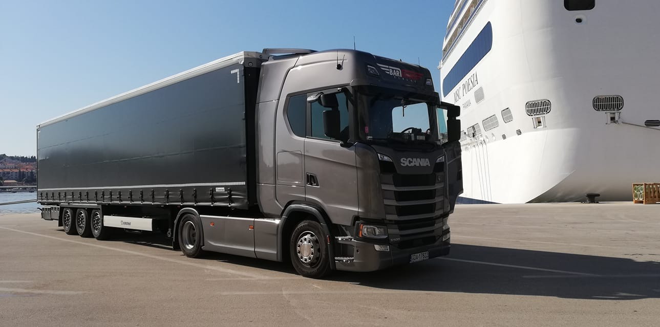 Szary ciągnik siodłowy Scania z naczepą na tle białego statku pasażerskiego MSC Poesia w porcie.