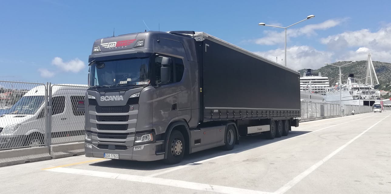 Szary ciągnik siodłowy Scania z naczepą na tle portu z widocznymi promami i elementami infrastruktury portowej, w słoneczny dzień.