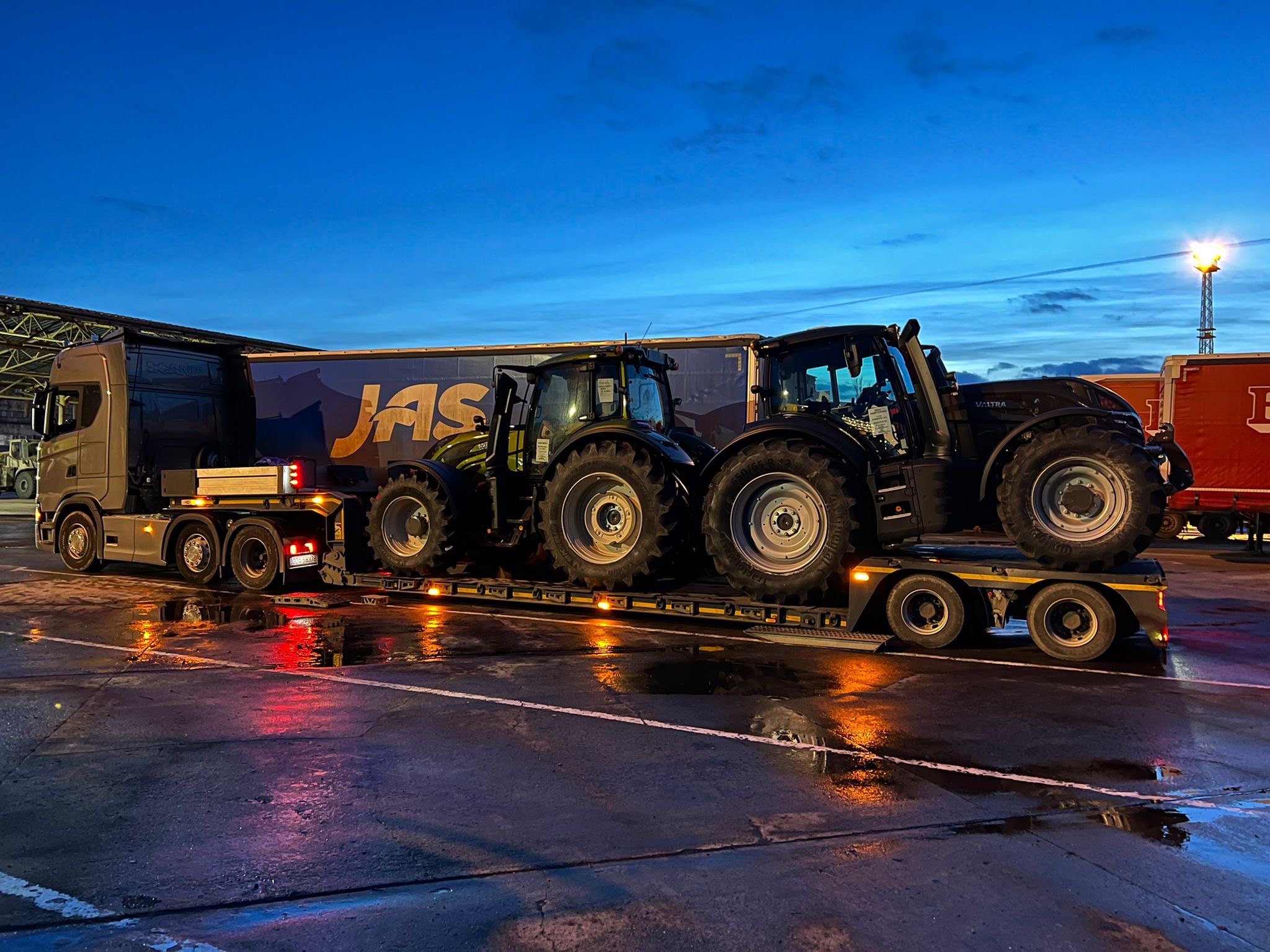 Ciężarówka z naczepą niskopodwoziową przewozi dwa czarne traktory Valtra na tle wieczornego nieba. Naczepa oznaczona logiem JAS.
