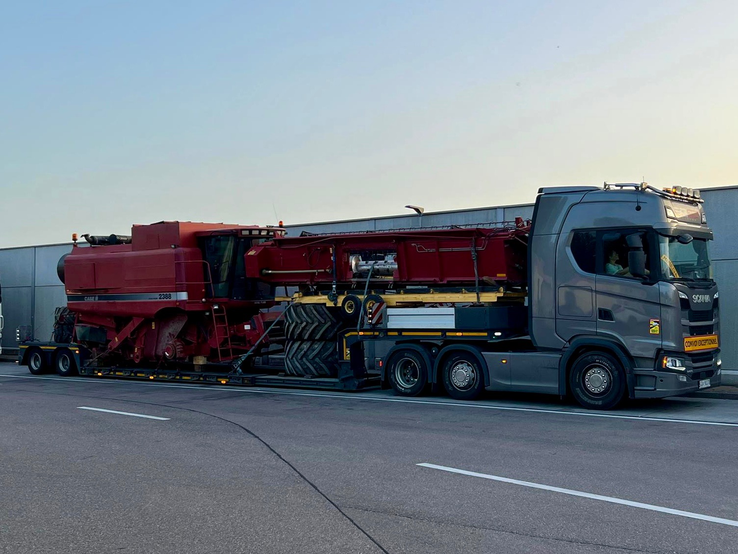 Szary ciągnik Scania z naczepą przewożącą kombajn rolniczy Case 2388 na lawecie z dodatkowymi oponami, tablica 'Convoi Exceptionnel'.