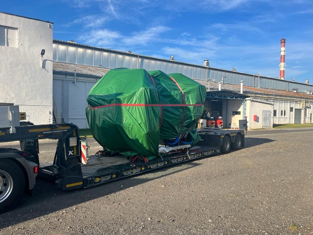 Nietypowy ładunek owinięty zieloną plandeką i zabezpieczony czerwonymi pasami, przewożony na niskopodwoziowej naczepie ciężarówki, w tle budynki przemysłowe i komin