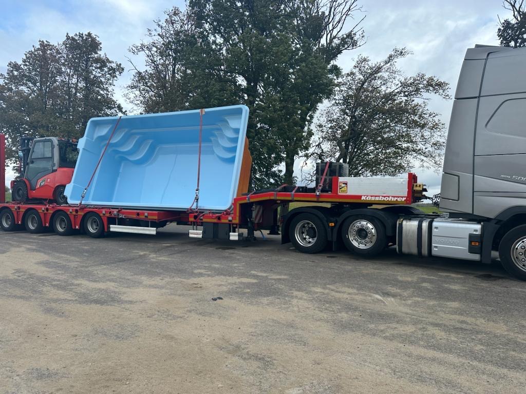 Niebieski, prefabrykowany basen z wyprofilowanymi schodami transportowany na niskopodwoziowej naczepie ciągniętej przez srebrną ciężarówkę Volvo, z widocznym w tle czerwonym wózkiem widłowym.