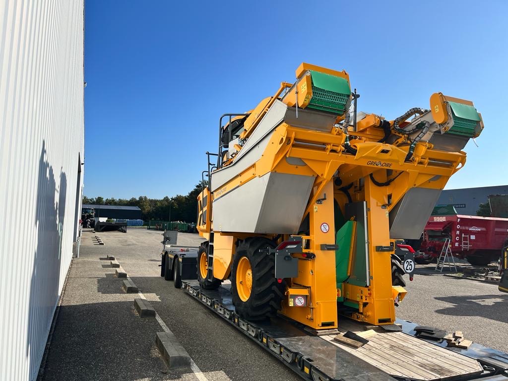 Żółta kombajn do zbioru winogron marki Grégoire, przewożony na niskopodwoziowej platformie transportowej na tle błękitnego nieba i zabudowań przemysłowych.
