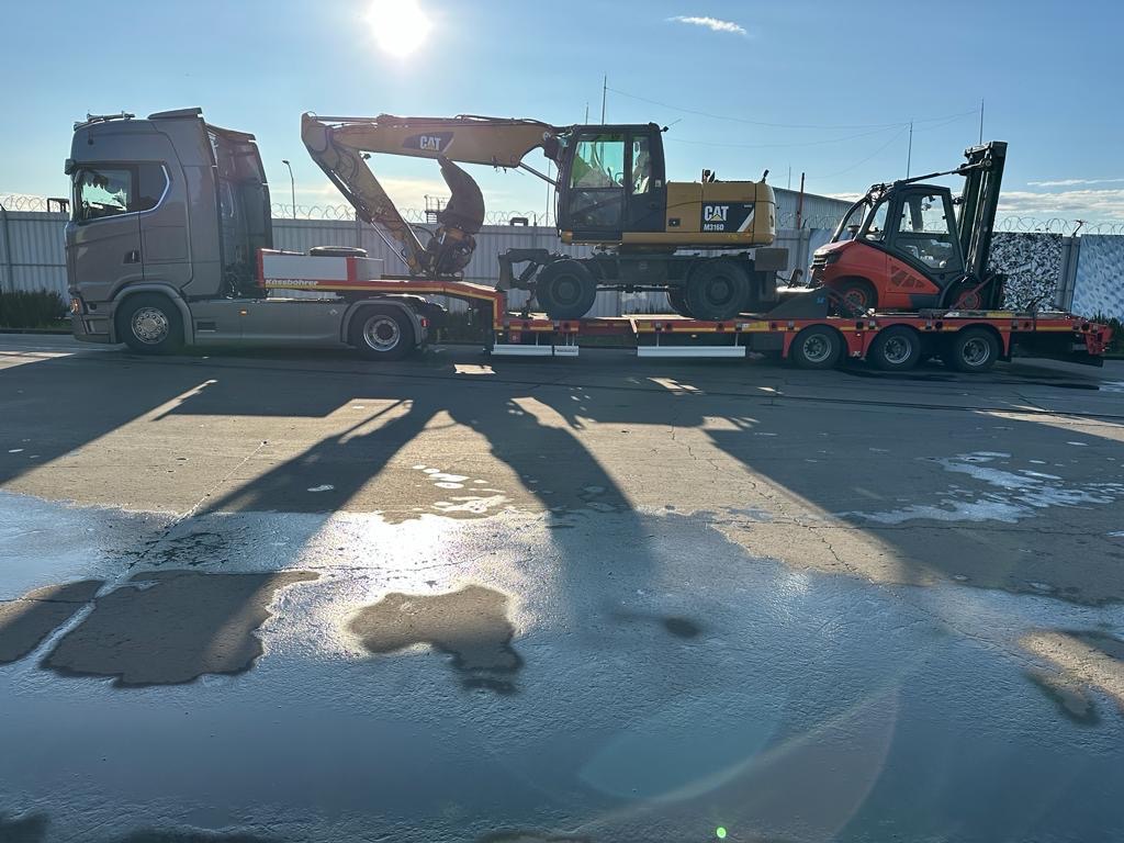 Ciężarówka z naczepą przewożąca koparkę CAT i wózek widłowy, zaparkowana na placu z mokrą nawierzchnią w słoneczny dzień.