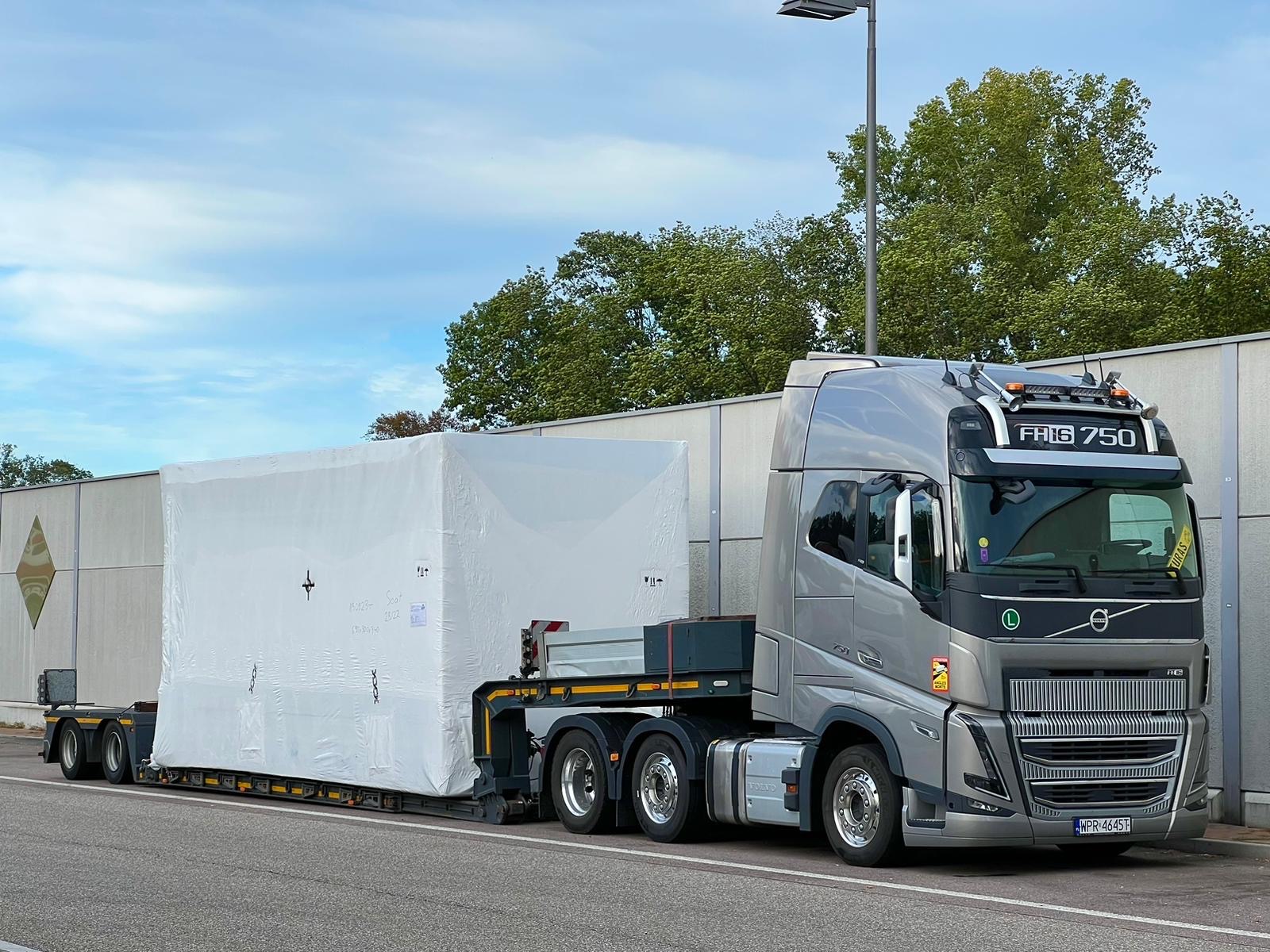 Srebrna ciężarówka Volvo FH16 750 przewożąca na niskopodwoziowej naczepie duży, biały ładunek zabezpieczony folią, na tle betonowego muru i drzew.