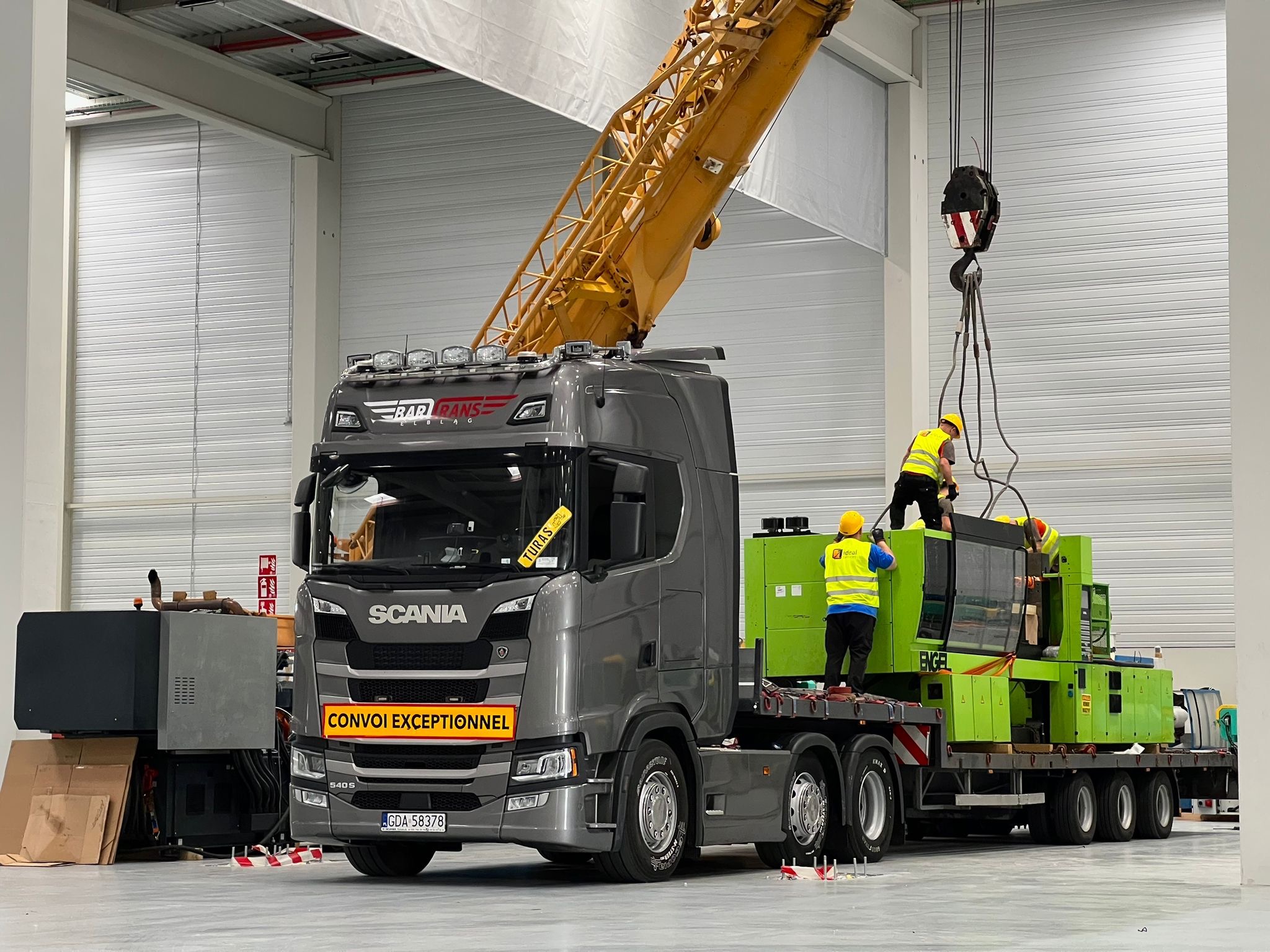Szary ciągnik siodłowy Scania z żółtym napisem 'CONVOI EXCEPTIONNEL' przewozi na platformie zieloną maszynę przemysłową, która jest podnoszona przez żółty dźwig w hali.