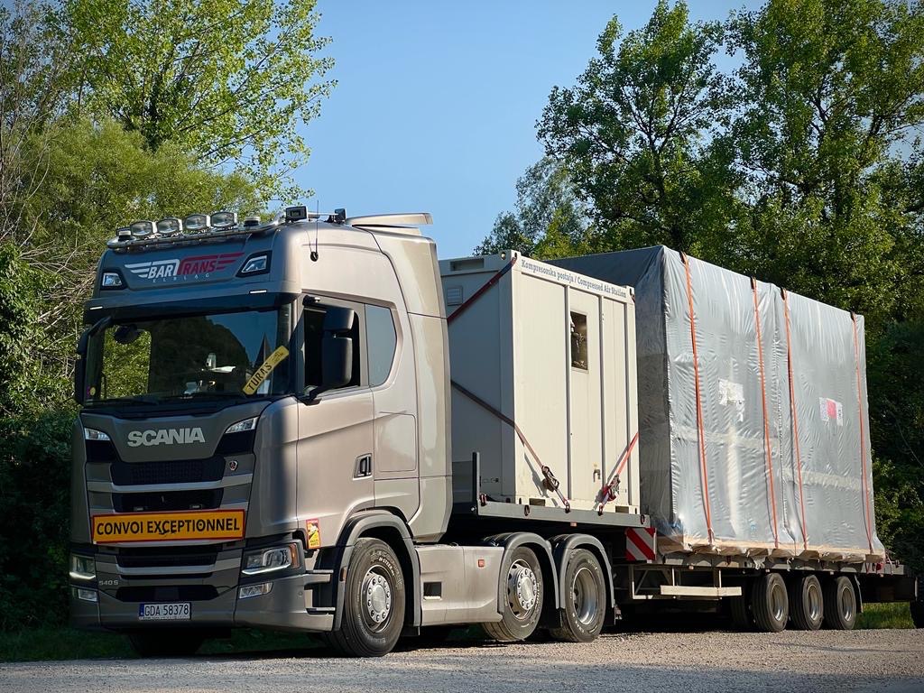 Szary ciągnik siodłowy Scania transportuje na niskopodwoziowej naczepie ładunek gabarytowy, częściowo zakryty srebrną plandeką, z tablicą 'CONVOI EXCEPTIONNEL'.