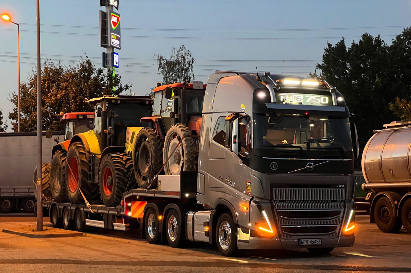 Szary ciągnik siodłowy Volvo FH16 750 przewozi dwa pomarańczowo-żółte traktory rolnicze na niskopodwoziowej platformie, widoczne światła pozycyjne i tablica rejestracyjna WPR 4645T.