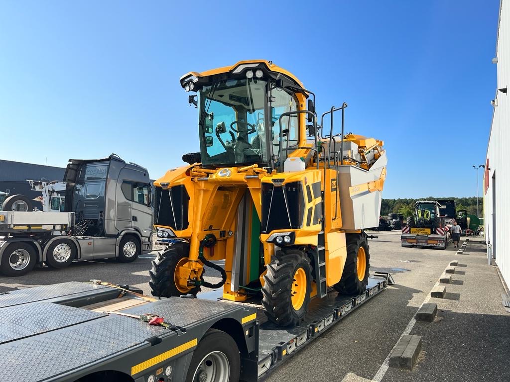 Żółty kombajn do zbioru winogron na niskopodwoziowej naczepie, z widoczną szarą kabiną ciężarówki w tle.