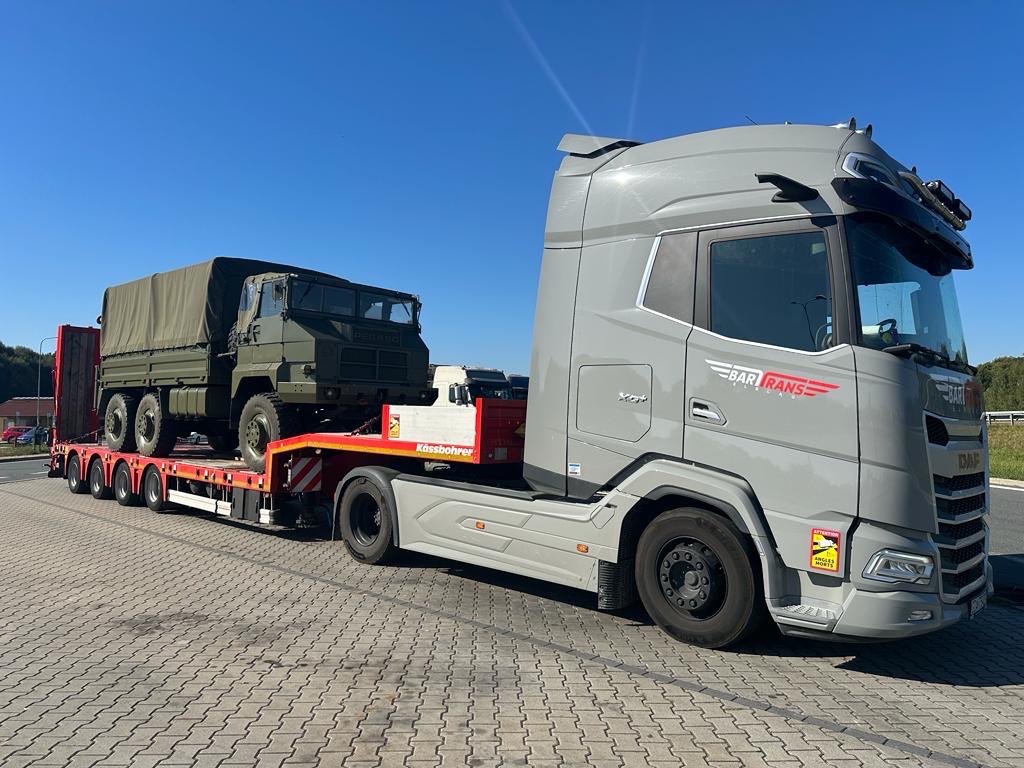 Szary ciągnik siodłowy z naczepą niskopodwoziową przewożącą wojskowy samochód ciężarowy z plandeką, na tle błękitnego nieba.